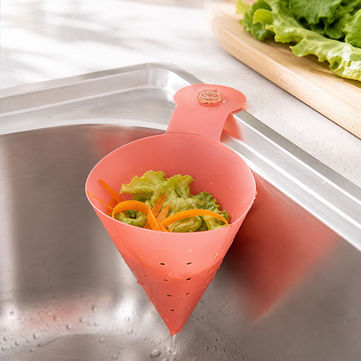 A pink silicone sink strainer with holes, holding lettuce and carrot peels, attached to a metal sink with water dripping.
