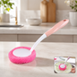 A pink and white long-handled dish scrubber with a round pink scrubbing pad rests on a kitchen counter.