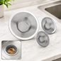 Three stainless steel mesh sink strainers of varying sizes on a white marble countertop next to a sink.