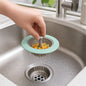 A hand holds a mint green and gray sink strainer filled with chopped vegetables under running water in a stainless steel
