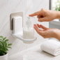A person dispenses white lotion from a clear bottle in a white wall-mounted holder onto their hand.