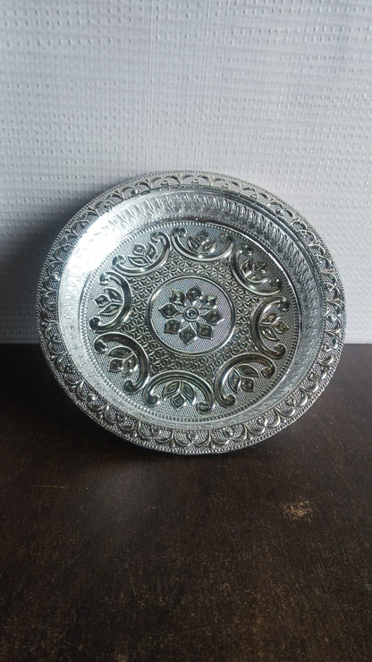 A silver-colored plastic pooja thali with embossed patterns, on a dark brown surface with a white textured wall in the