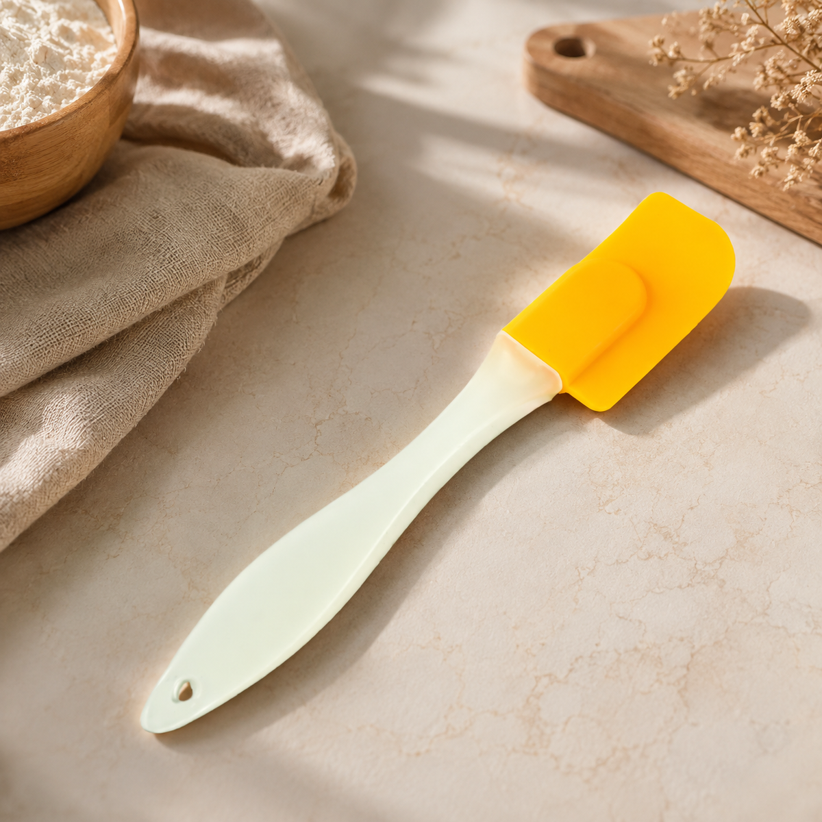 A yellow silicone spatula with a light cream handle rests on a marble counter next to a bowl of flour and a beige cloth.