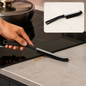 A hand uses a black and gray crevice cleaning brush to sweep crumbs off a kitchen counter next to a stovetop.