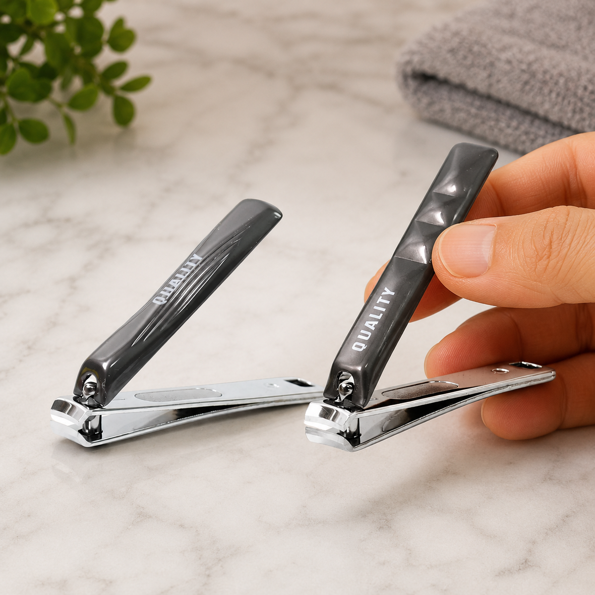 A hand holds one of two black and silver nail clippers on a marble surface with a green plant and grey towel.