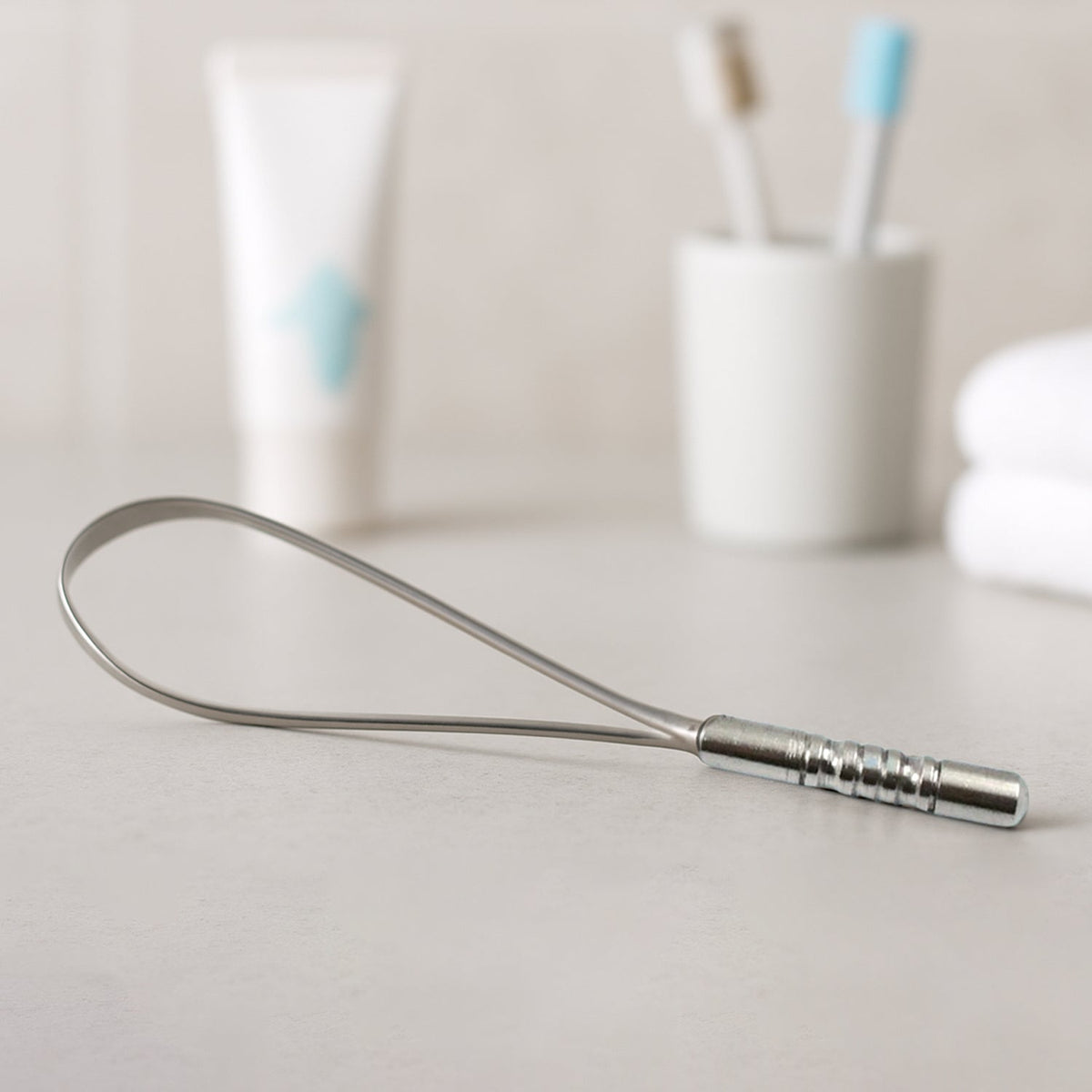 A silver metal tongue cleaner rests on a light gray countertop, with blurred toiletries in the background.