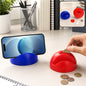A blue phone holder with a phone and a red piggy bank with coins being inserted on a white desk.