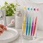 A hand holds a red toothbrush with toothpaste over a sink, while five other colorful toothbrushes stand in a glass.