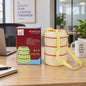A cream-colored three-layer lunch box with a yellow strap sits next to its red and green packaging on a wooden desk.