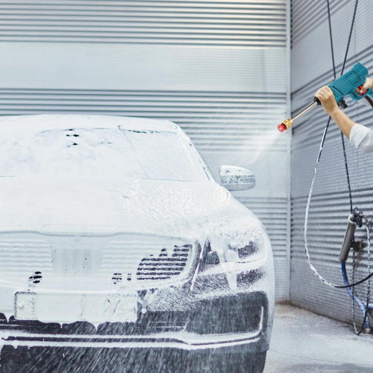 A person's hands hold a teal pressure washer, spraying water onto a white foamy car in front of a gray garage door.