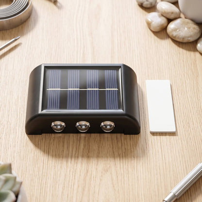 A black solar-powered LED wall light with a solar panel and three sensors, next to a white adhesive strip on a wooden table