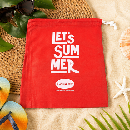 A red drawstring bag with "LET'S SUMMER" and Havaianas logo, surrounded by sunglasses, flip-flops, and beach decor on sand.