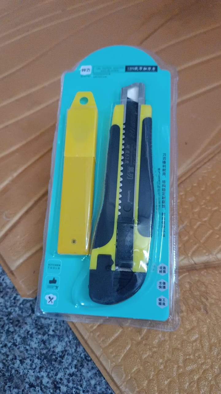 A yellow and black utility knife with its yellow blade cover, sealed in clear retail packaging.
