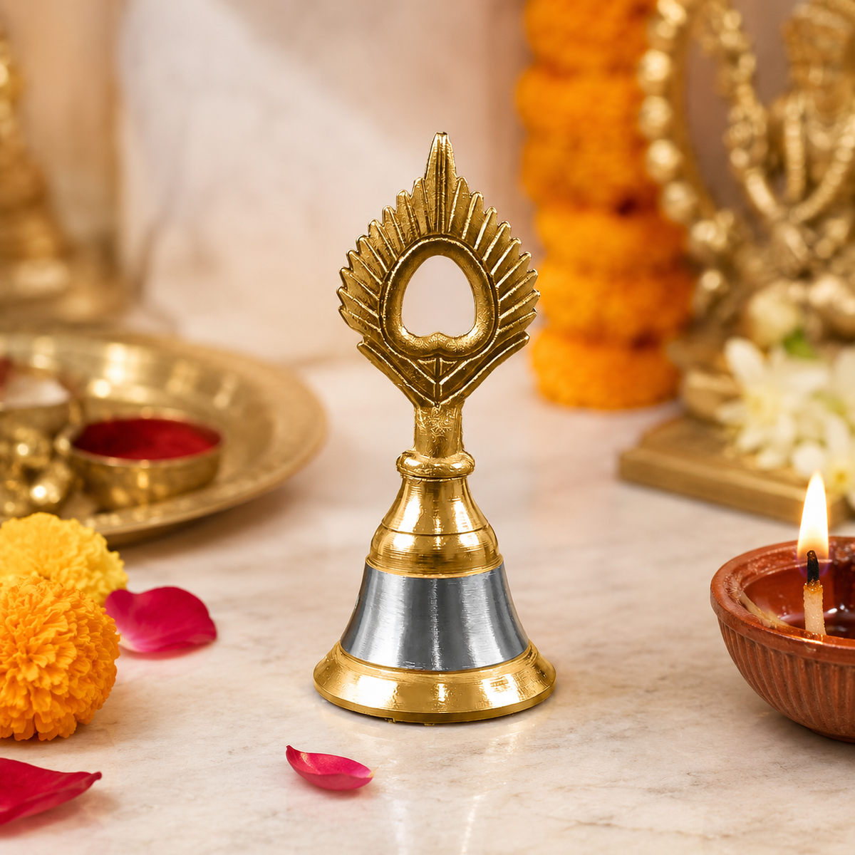 A small brass and steel pooja bell with a peacock feather design sits on a marble surface amidst marigolds and lit diyas.
