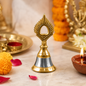 A small brass and steel pooja bell with a peacock feather design sits on a marble surface amidst marigolds and lit diyas.