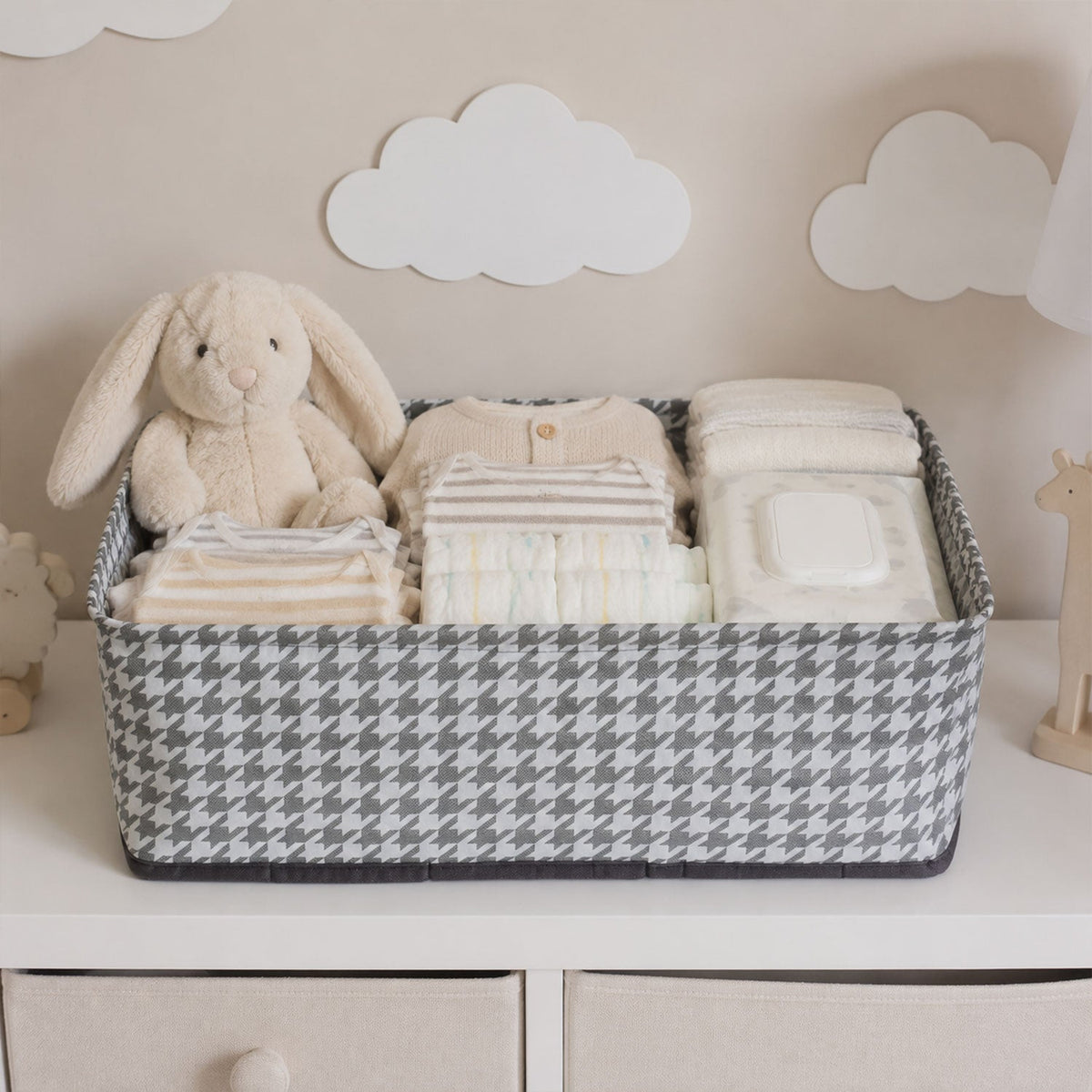 A gray and white houndstooth fabric basket holds baby clothes, diapers, wipes, and a plush bunny on a white dresser.
