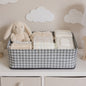A gray and white houndstooth fabric basket holds baby clothes, diapers, wipes, and a plush bunny on a white dresser.