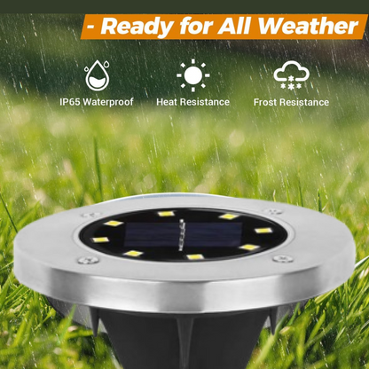 A close-up of a silver solar ground light with a black solar panel and eight yellow LED lights, surrounded by green grass.