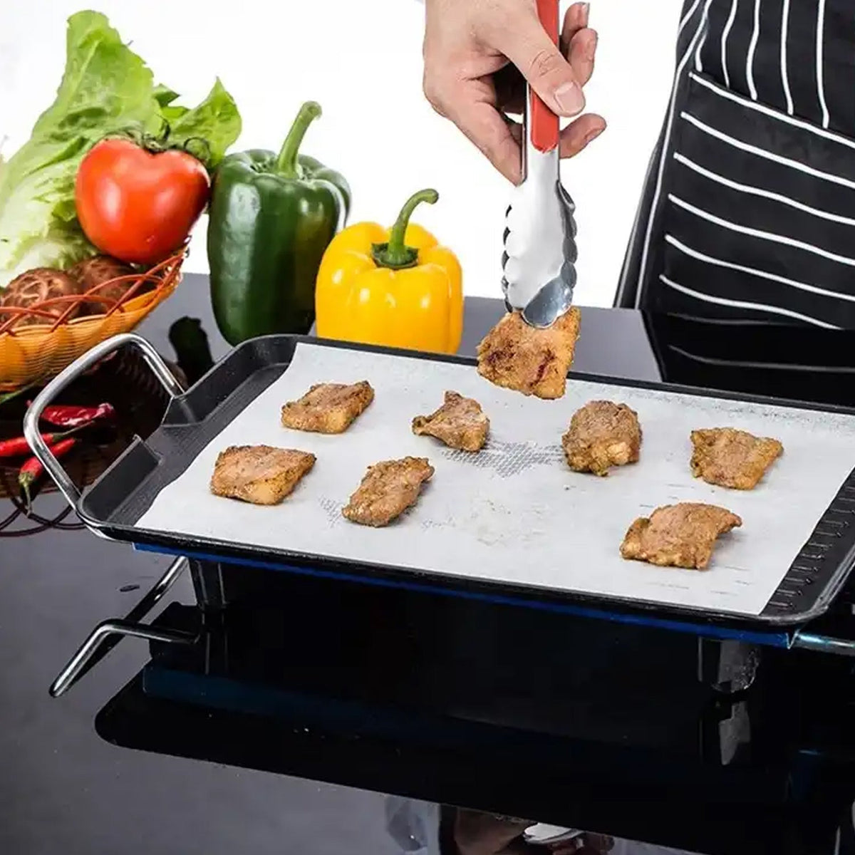 A hand using red tongs to place a piece of cooked meat onto a baking sheet lined with non-stick paper, surrounded by fresh