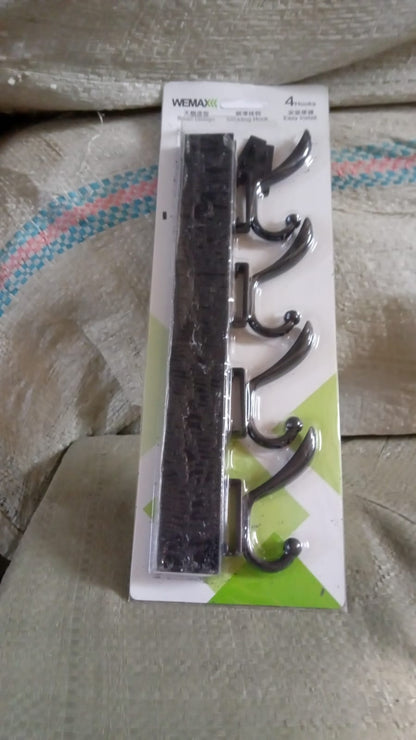 Black four-hook coat rack in its clear plastic packaging with a white and green label, resting on a textured surface.