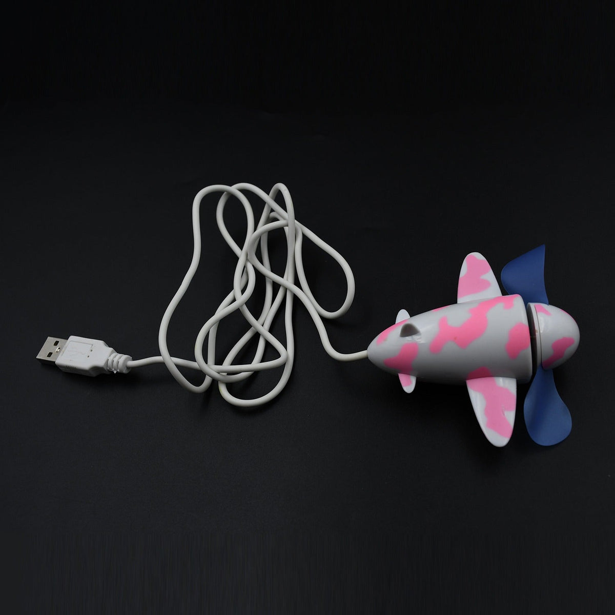 A pink and white airplane-shaped USB fan with blue propellers and a coiled white USB cable on a dark surface.