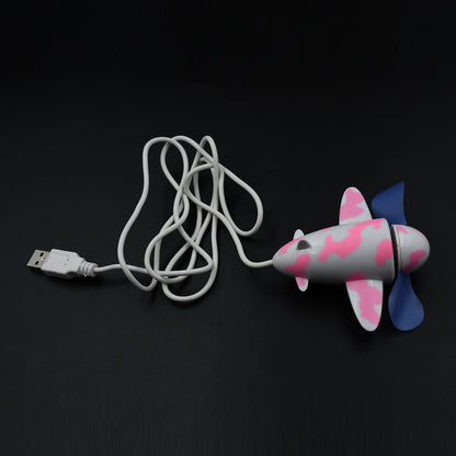 A pink and white airplane-shaped USB fan with blue propellers and a coiled white USB cable on a dark surface.