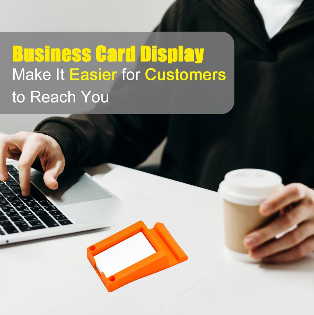 An orange plastic holder with white business cards on a white desk, with a person typing on a laptop in the background.