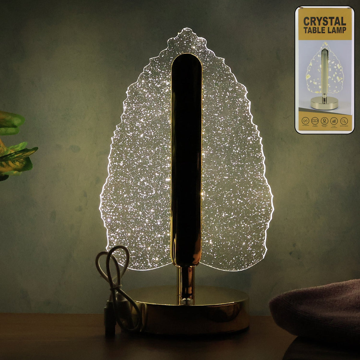 Glowing crystal leaf lamp with gold base and stem on a dark wooden surface, with product packaging.