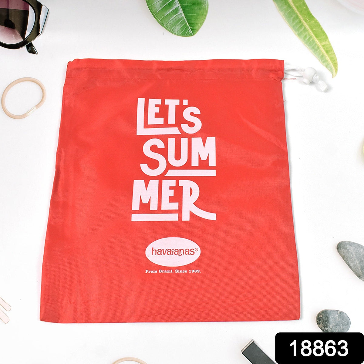 A red drawstring bag with "LET'S SUMMER" and Havaianas logo, on a white surface with green leaves and small grey stones.
