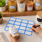 A hand writes 'RICE' on a blue-bordered label sticker on a jar, with other labeled jars in the background.