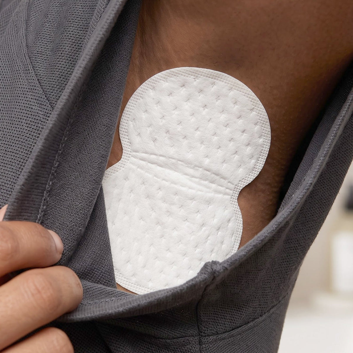 A white sweat pad is shown adhered to the armpit of a person wearing a grey t-shirt, with a hand adjusting the shirt.