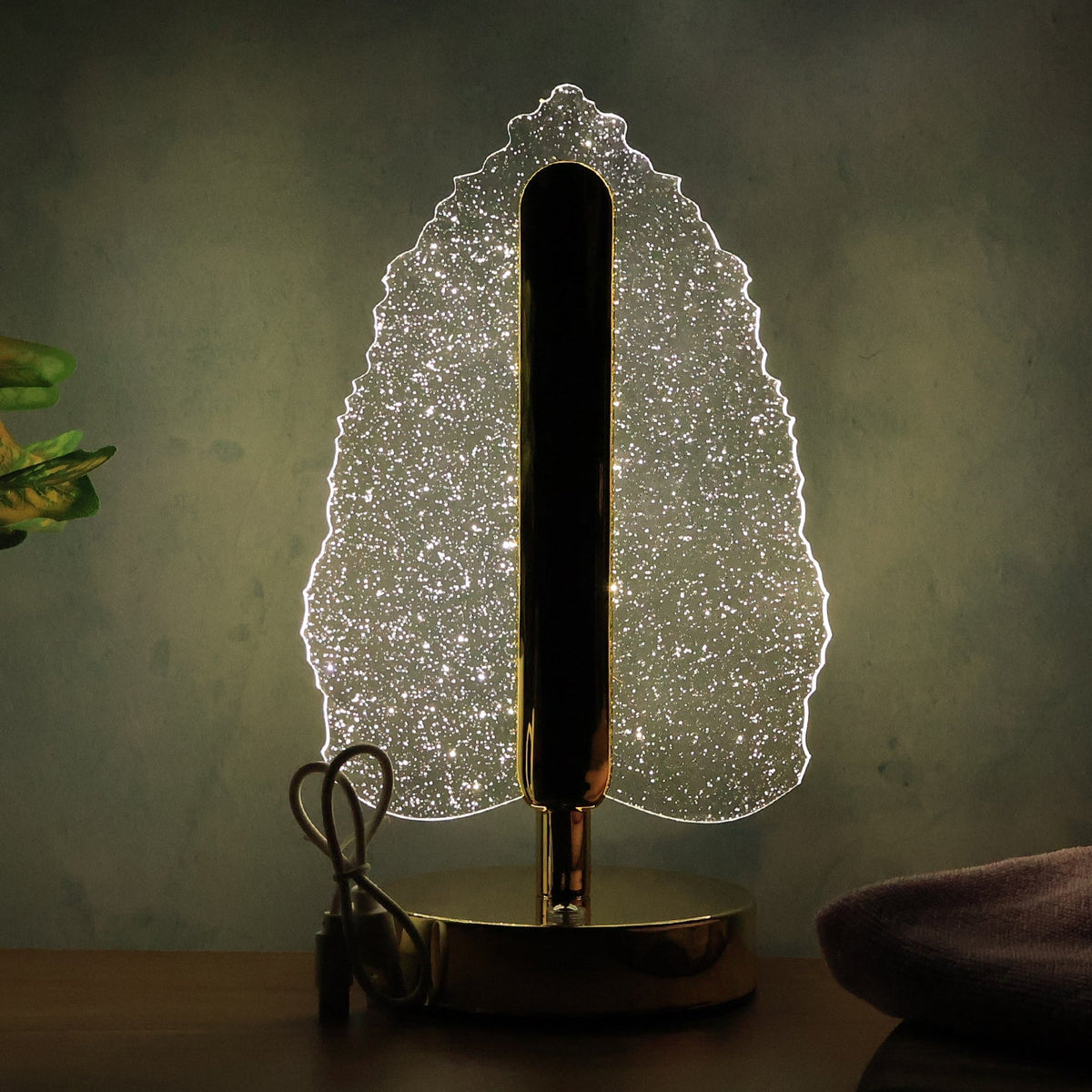 Crystal leaf lamp with gold base and stem, glowing softly on a dark wooden surface with a plant nearby.