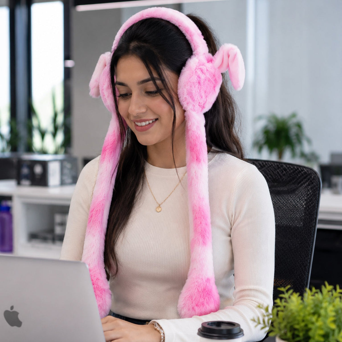 A young woman smiling and working on a laptop while wearing pink and white plush bunny earmuffs with long tails in an office