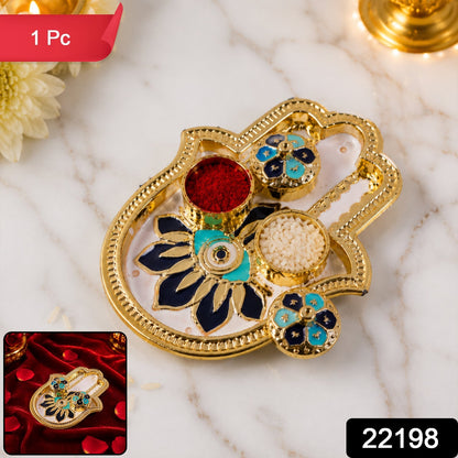 Golden hamsa hand thali with red kumkum, white rice, and blue and black enamel floral designs.