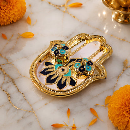 Golden hamsa hand thali with blue and black enamel, on a marble surface with scattered orange petals and flowers.