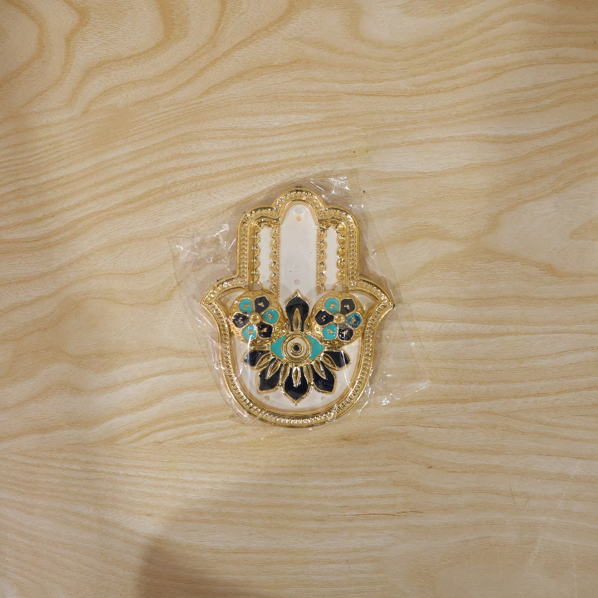Golden hamsa hand thali with blue and black enamel details, packaged in clear plastic on a wooden surface.