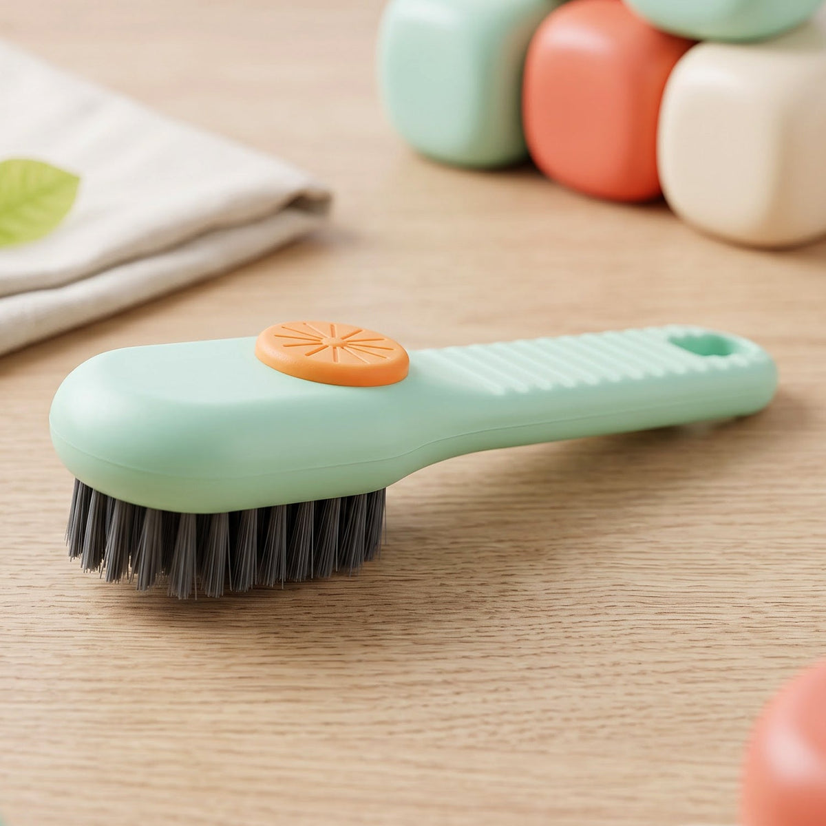 A green and orange liquid-dispensing brush rests on a wooden surface, with blurred colorful containers in the background.