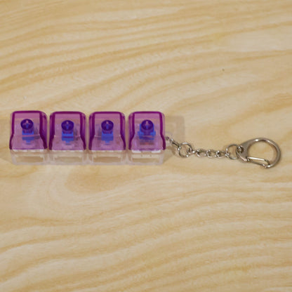 A four-key keyboard switch keychain with purple caps, clear bases, and a metal clasp on a light wooden surface.