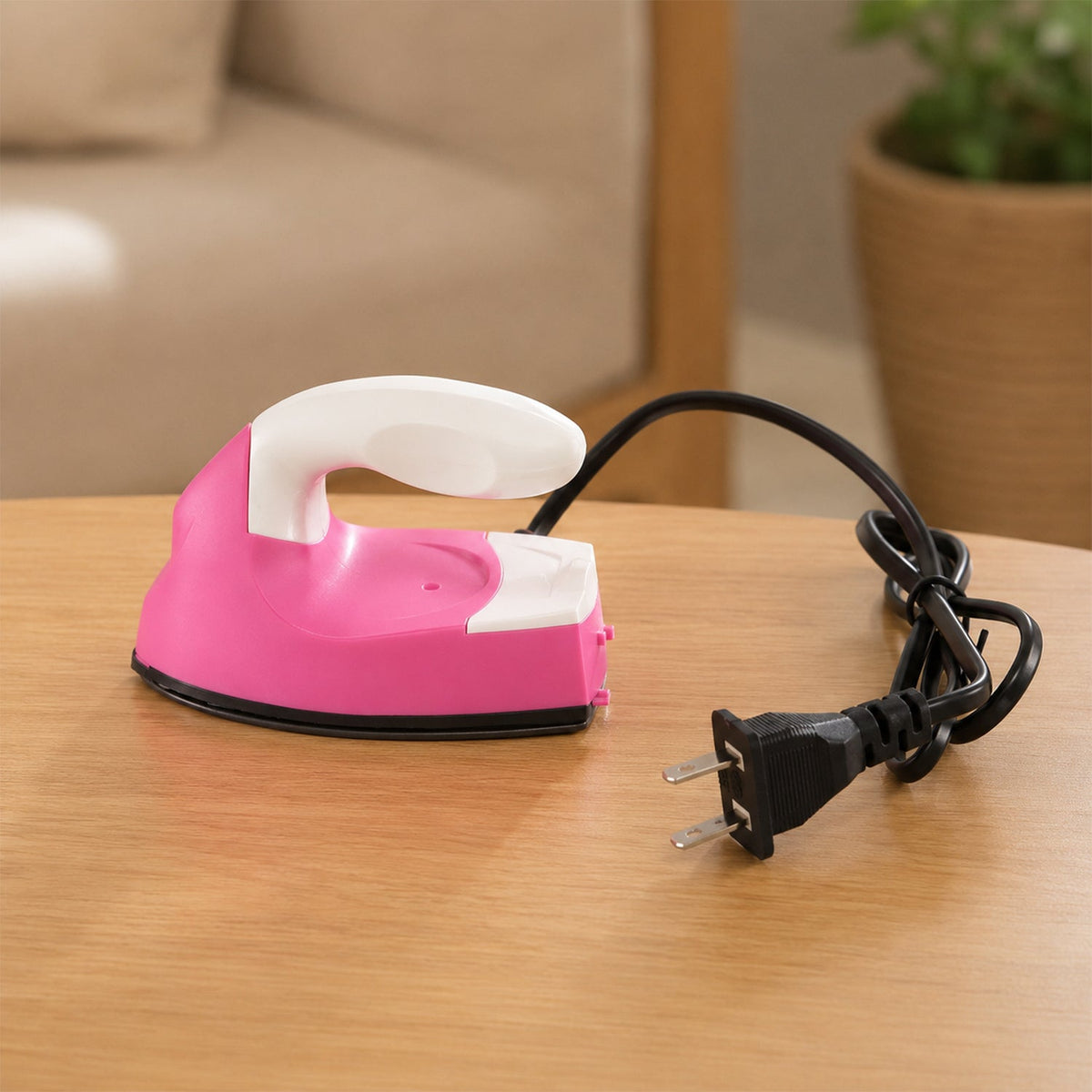 A pink and white mini iron with its black power cord and plug rests on a wooden table in a room.