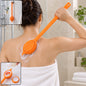 A person uses an orange silicone back scrubber with a soap dispenser on their back, creating suds.