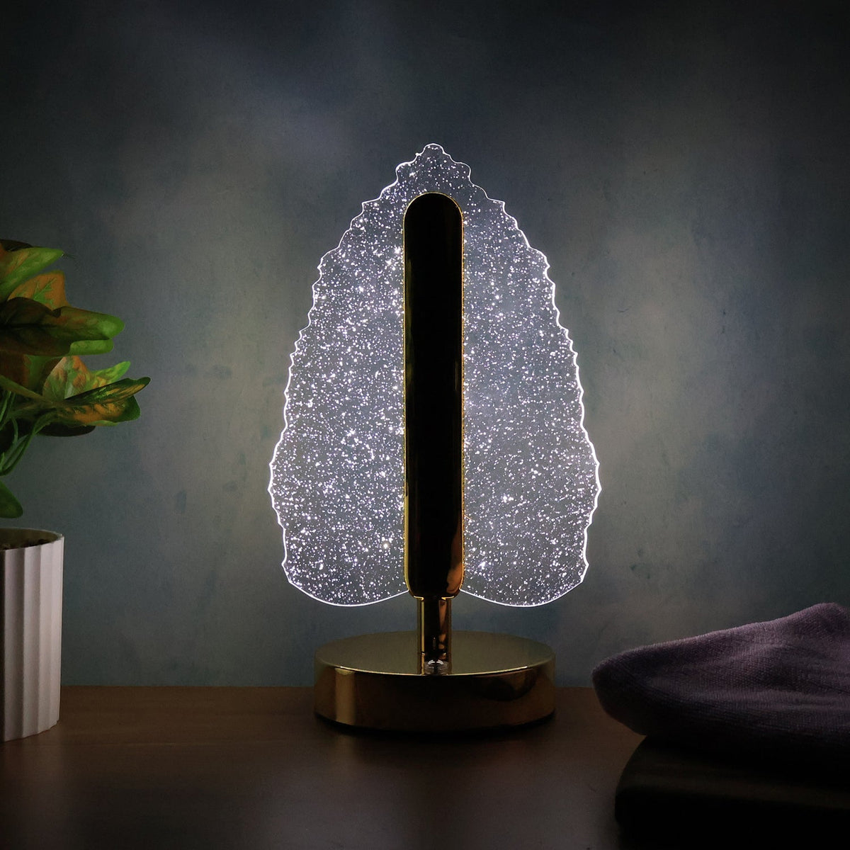 Illuminated crystal leaf lamp with a gold base and stem on a dark wooden surface with a plant.