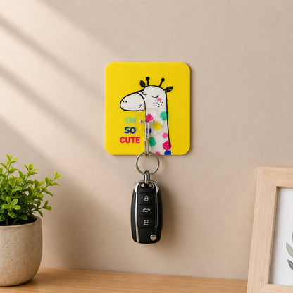 A yellow square wall hook with a cartoon giraffe and text "I'M SO CUTE" holds a black car key, with a potted plant and