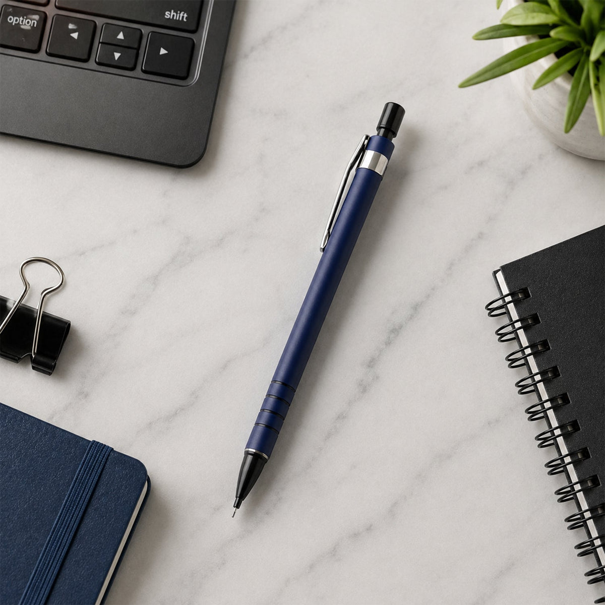 A blue mechanical pencil lies on a white marble desk surrounded by a black notebook, a laptop keyboard, and a small plant.
