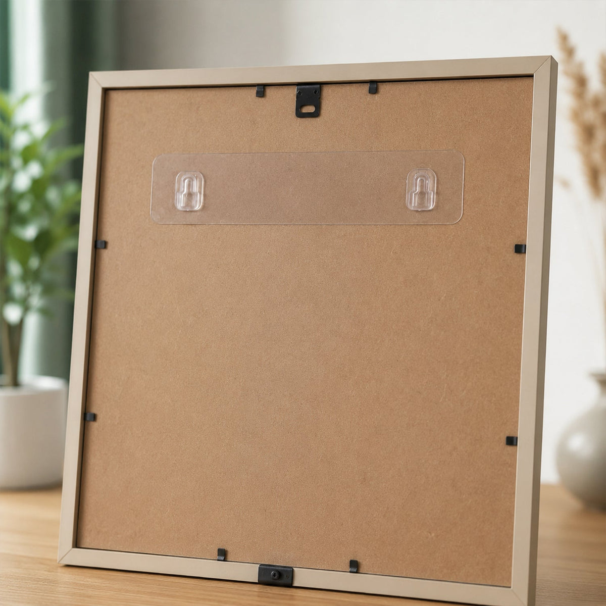 A clear adhesive wall hook strip applied to the back of a light brown picture frame, with a plant in the background.