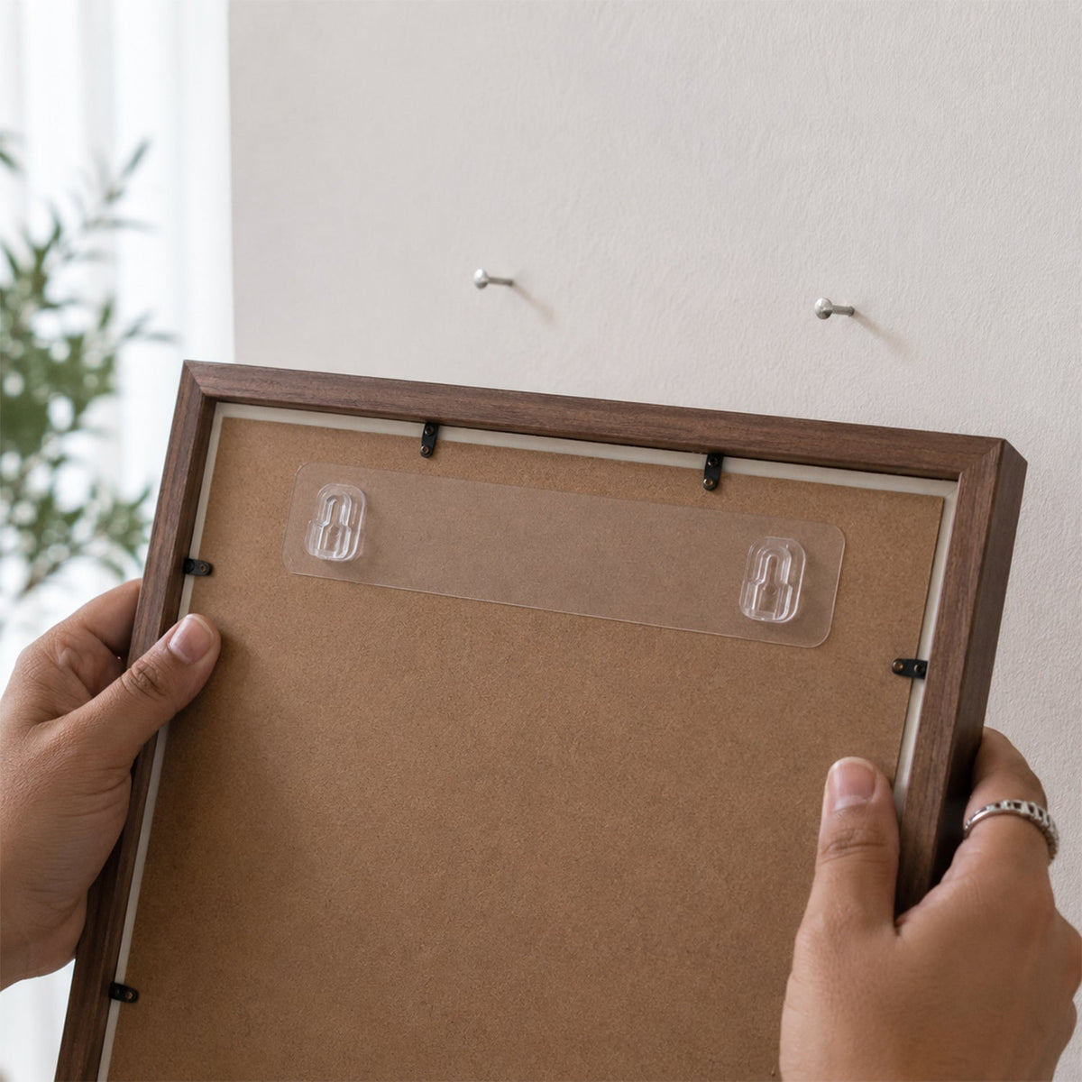 Hands holding a brown picture frame with an applied clear adhesive hook strip, about to hang it on a white wall with two