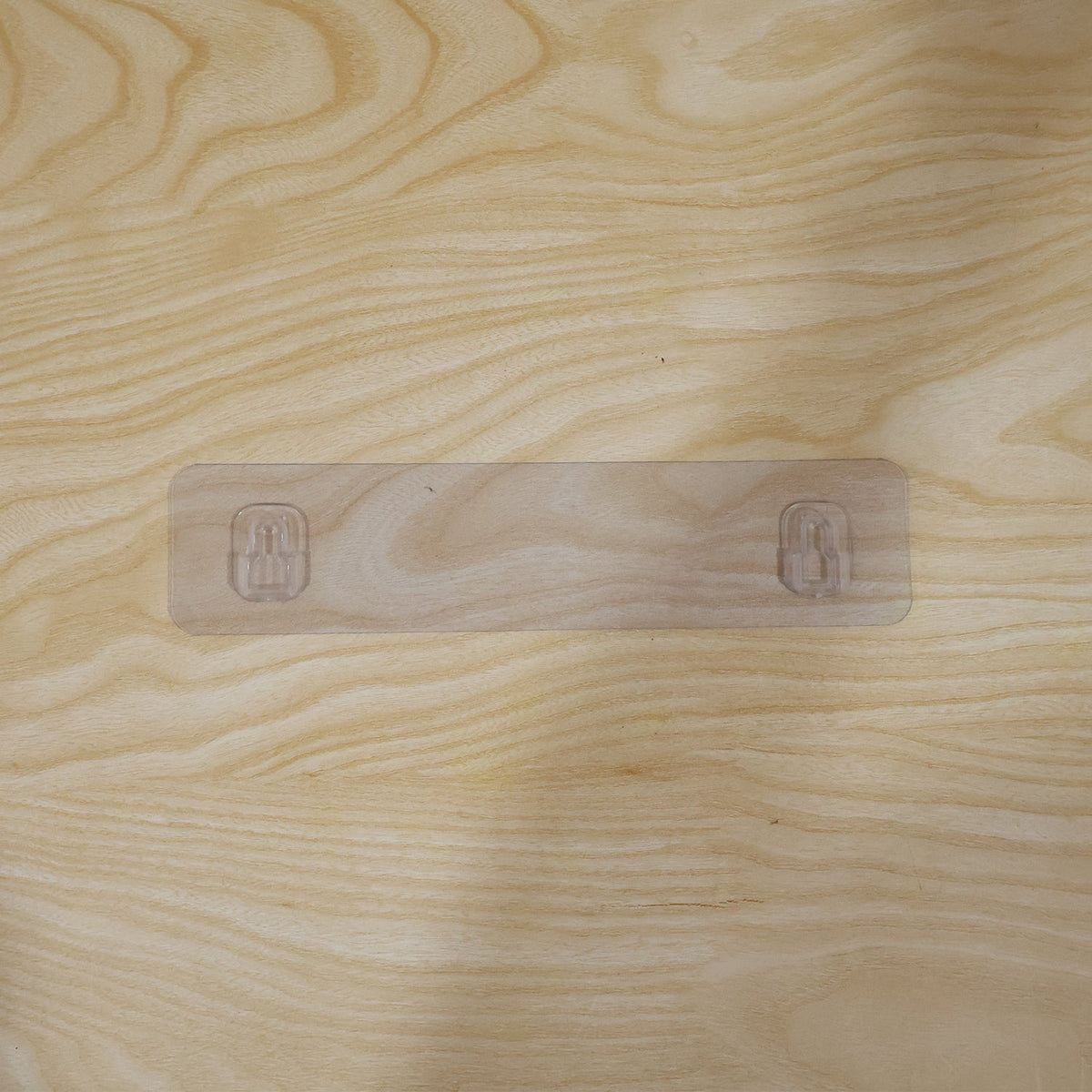 A clear adhesive wall hook strip with two hooks applied to a light wooden surface.