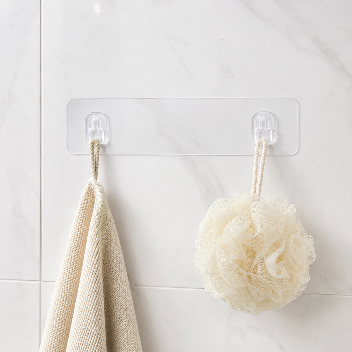 A clear adhesive wall hook strip with two hooks, holding a beige towel and a cream bath pouf against a white tiled wall.