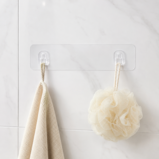 A clear adhesive wall hook strip with two hooks, holding a beige towel and a cream bath pouf against a white tiled wall.