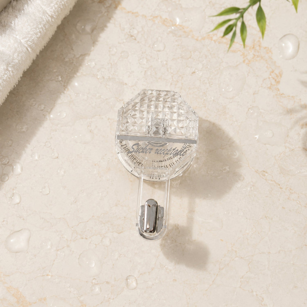 A clear, diamond-patterned self-adhesive hook with a silver hook on a light marble surface with water droplets.