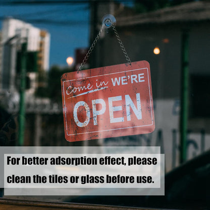 A red 'Come in We're Open' sign hangs by a chain from a suction cup hook on a glass window, with blurred buildings outside.